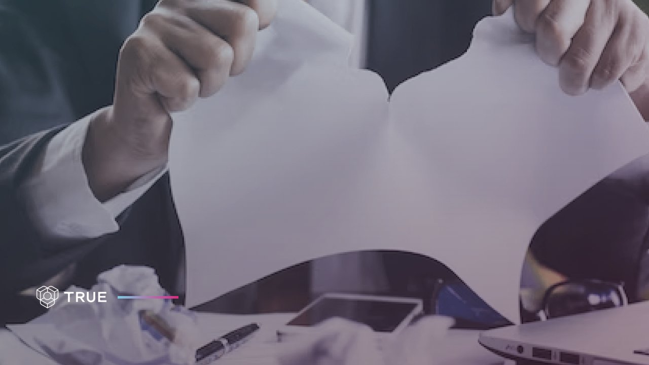 Document fraud: a man in a suit tears a paper document in two over a desk with crumpled paper, a pen and a laptop